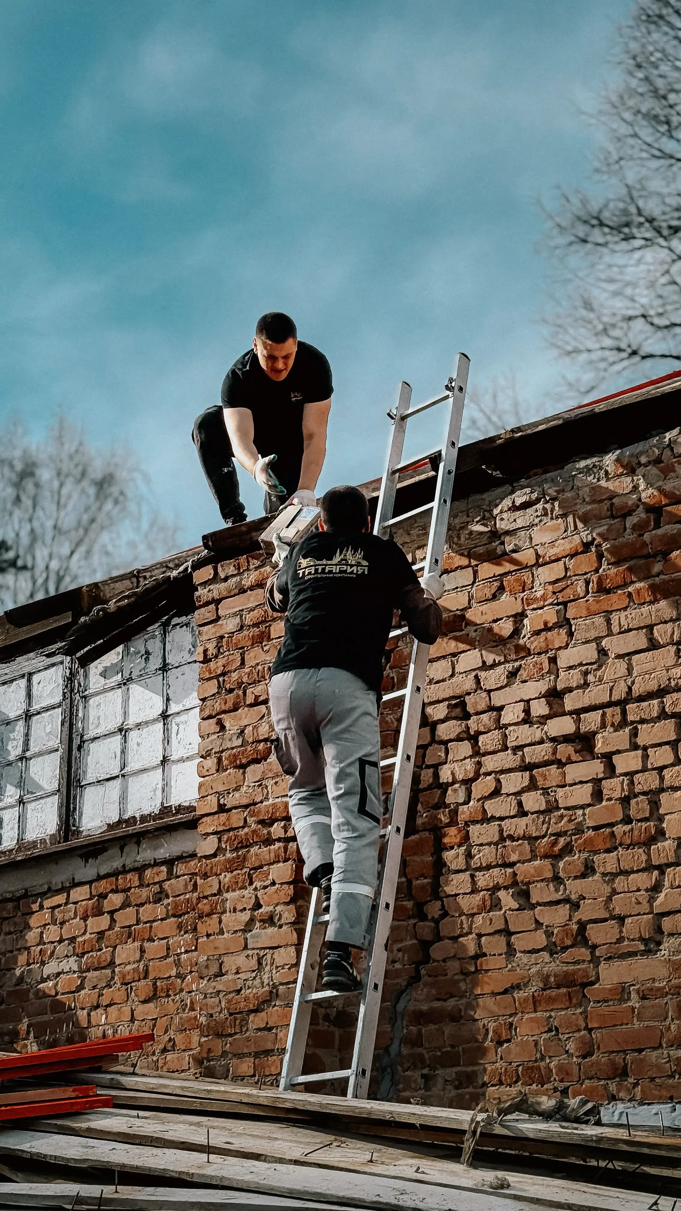 Zwei Handwerker reichen sich Material über eine Leiter an einer Backsteinfassade